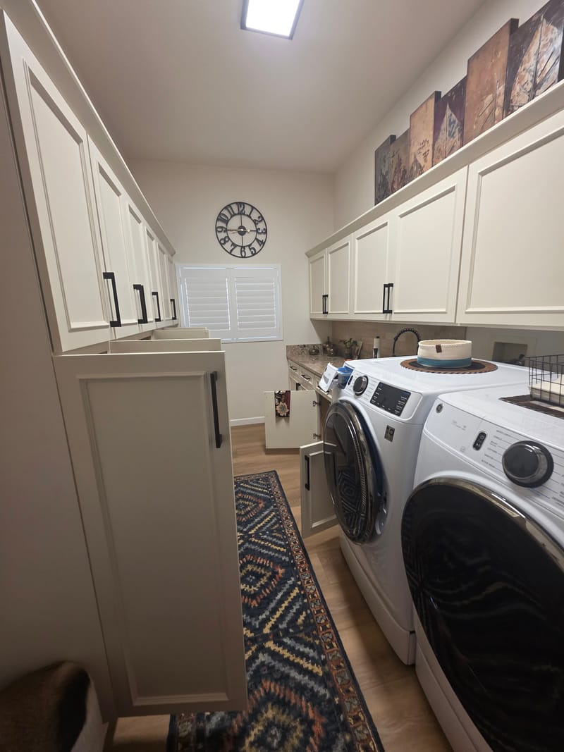Narrow laundry room with white cabinets on both walls, a front loading washer and dryer, and a patterned runner on the floor